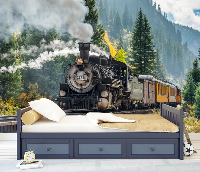 May include: A dark blue wooden bed frame with three storage drawers underneath, featuring a white mattress, a white pillow, a cylindrical yellow bolster, and a tan blanket. The bed is positioned against a wall mural of a steam locomotive.