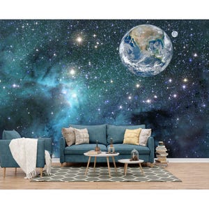 May include: A teal sofa and armchair set with throw pillows and a white blanket. The furniture sits on a patterned rug in front of a wall mural depicting Earth, the moon, and a starry galaxy. Two small tables and a stack of books complete the room.
