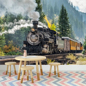 May include: A round light-coloured wooden table with three matching stools sits on a chevron-patterned rug. The wall behind features a large mural of a black steam locomotive numbered 482 travelling on tracks through a forest with mountains.