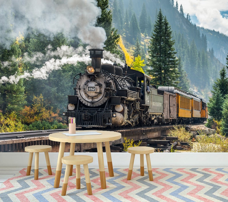 May include: A round light-coloured wooden table with three matching stools sits on a chevron-patterned rug. The wall behind features a large mural of a black steam locomotive numbered 482 travelling on tracks through a forest with mountains.