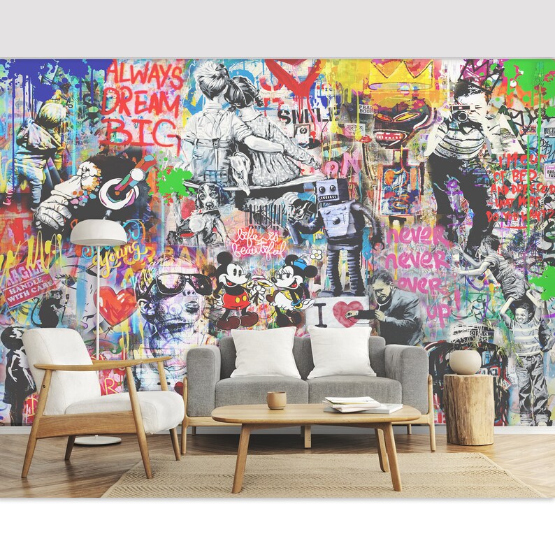 Op de afbeelding: Een grote muurschildering met een levendige graffiti-stijl collage met kleurrijke verfspatten, stripfiguren zoals Mickey en Minnie Mouse, een robot en tekst waaronder "ALWAYS DREAM BIG" en "never never over up" op een witte muur.