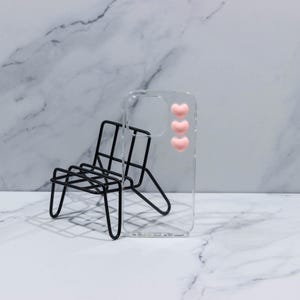 Può includere: Una custodia trasparente per telefono con tre decorazioni a forma di cuore rosa. Una piccola sedia in metallo nero è posizionata davanti alla custodia. Lo sfondo presenta un motivo marmorizzato bianco e grigio.