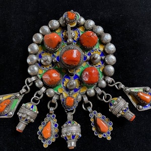 May include: A silver pendant with a round, enamel-painted center featuring red coral stones and yellow, green, and blue accents. The pendant has multiple dangling charms with red coral stones and silver beads.
