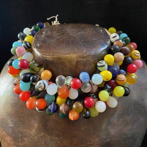 Può includere: Una collana a più fili composta da numerose piccole perle di vetro a forma di goccia in un'ampia varietà di colori, tra cui rosso, giallo, blu, verde, arancione, bianco e nero. Alcune perle presentano motivi a vortice, altre sono solide o traslucide.