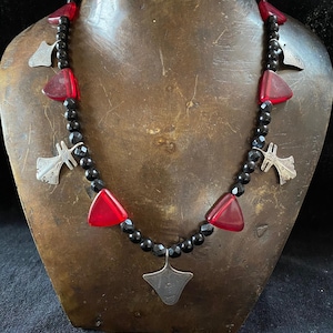 May include: A black beaded necklace with red glass triangle beads and silver pendant charms. The necklace is displayed on a brown mannequin.