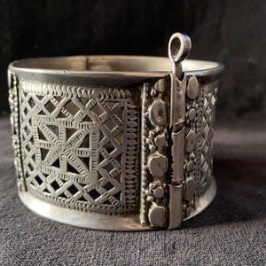 May include: Silver-toned cuff bracelet with intricate openwork geometric patterns. The hinged design features decorative rivets and a small loop closure. The bracelet's surface has a slightly aged appearance, suggesting it is vintage or antique.