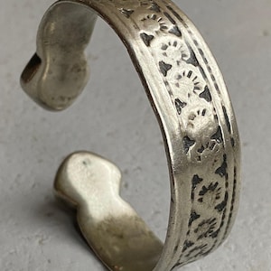 May include: A silver bracelet featuring a decorative floral pattern. The bracelet is crafted from thick, solid metal and has a simple, elegant design.