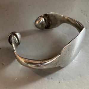 May include: A silver-toned cuff bracelet with a rounded, bulbous end and a decorative, textured design. The bracelet has a curved, open design and appears to be made of metal. The cuff is resting on a light gray surface.