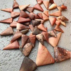 Puede incluir: Una colección de cuentas de piedras preciosas de forma triangular, de color rojo, marrón y naranja. Las cuentas están enhebradas en un alambre fino y están listas para convertirse en joyas.