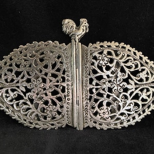 May include: Silver metal belt buckle with an elaborate openwork design of flowers and leaves. A small rooster figure is at the top. The buckle is oval and has a hinged closure. The buckle is approximately 10 cm in length.