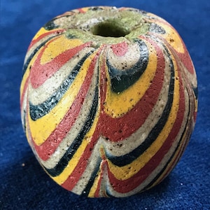 Könnte beinhalten: Eine farbenfrohe, runde Perle mit einem Loch in der Mitte. Die Perle weist wirbelnde Muster aus Rot, Gelb, Schwarz und Weiß auf. Die Oberseite der Perle ist in einem gedämpften Grünton gehalten.
