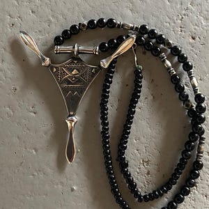 May include: A silver pendant with an intricate geometric design, paired with a necklace of black beads. The pendant has a unique shape with two wing-like extensions. The necklace is composed of small, round beads.
