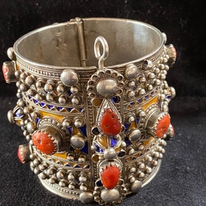 May include: A silver bracelet with intricate details, featuring a geometric design with blue and yellow enamel and red coral stones.