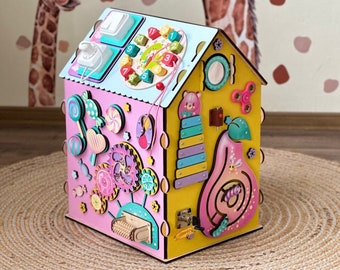 May include: A colorful wooden busy board shaped like a house. The pink and yellow house features various interactive elements, including gears, a clock, a light switch, and a xylophone. Decorative candies and a pear shape are also included.