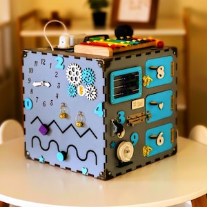 May include: A grey wooden activity cube with interactive features. It includes a clock, gears, abacus, latches, and a xylophone. The cube has blue and white accents, designed for early childhood development and learning.