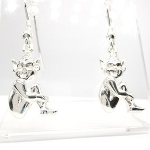 May include: A pair of silver earrings featuring a detailed sculpture of a sitting fairy with wings. The earrings are on a white display stand.
