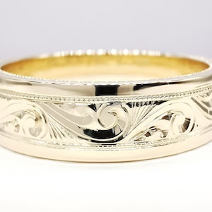 May include: A polished gold band ring featuring an intricate engraved scrollwork pattern that wraps around the exterior. The design is bordered by fine milgrain detailing along the top and bottom edges, creating a textured finish on the metal.