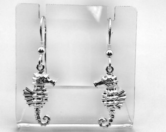 May include: Silver dangle earrings featuring a detailed seahorse design. The earrings are hanging from a clear display stand.
