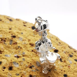 May include: Silver charm of a person playing a drum, wearing a hat. The charm is on a textured yellow surface.