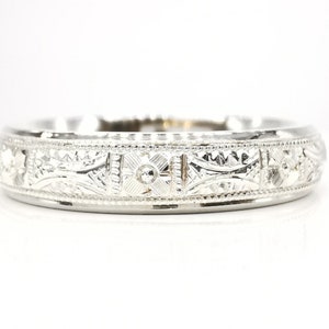 May include: A silver wedding band with an intricate engraved design. The band has a delicate floral pattern and a small diamond in the center.