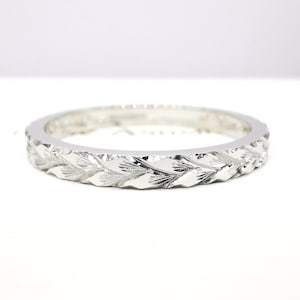 May include: A silver ring with a repeating leaf pattern. The ring is circular and has a textured surface with a series of detailed leaf designs around the band. The ring is set against a white background.