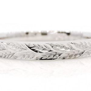 May include: A silver-toned metal ring featuring a continuous engraved pattern of small, overlapping leaves around the entire outer band. The surface of the ring has a polished, reflective finish that catches the light, highlighting the intricate details of the botanical-inspired carvings etched into the metal.