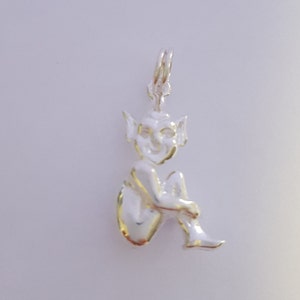 May include: A silver charm in the shape of a seated goblin with pointed ears and a smiling face. The goblin is in a relaxed pose, with its legs bent and arms wrapped around its knees. The charm has a loop at the top for attaching to a necklace or bracelet.