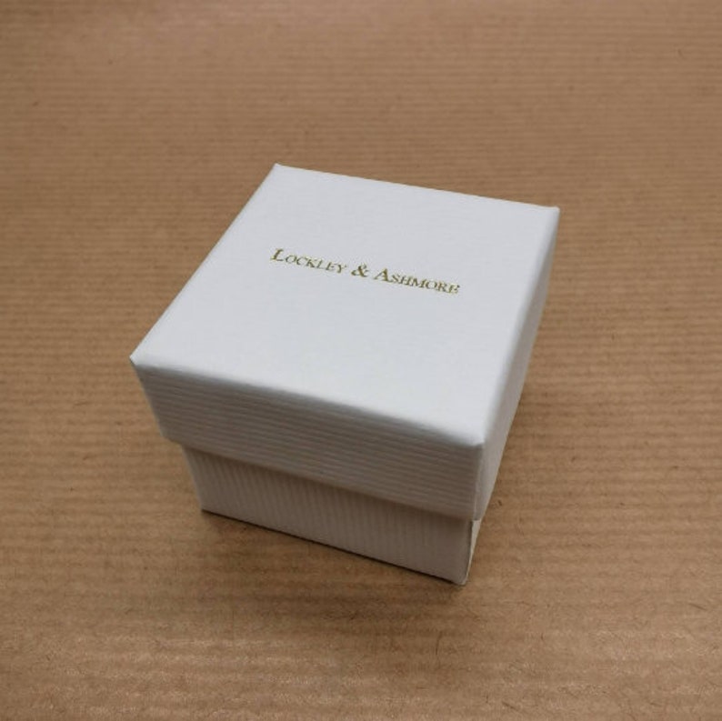 Puede incluir: Caja cuadrada blanca con el texto "Lockley & Ashmore" impreso en letras doradas.
