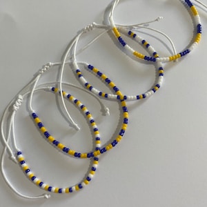 May include: A set of four adjustable bracelets featuring a repeating pattern of small white, yellow, and royal blue beads strung on thin white cord. Each bracelet includes a sliding knot closure mechanism to allow for a customisable fit on the wrist.