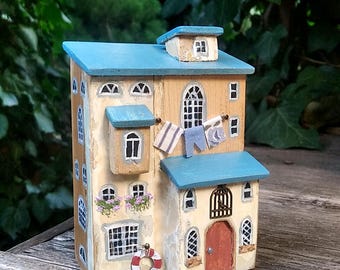 May include: A decorative wooden house figurine featuring a cream-colored facade, a blue roof, and multiple windows. The exterior includes painted details like a red and white life ring, hanging laundry, flower boxes, and a small arched brown door.