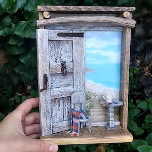 May include: A miniature wooden diorama featuring a weathered gray door with a metal latch and knocker. Inside, a small chair with a plaid blanket sits next to a round table holding a white cup. The scene opens to a painted coastal beach view.