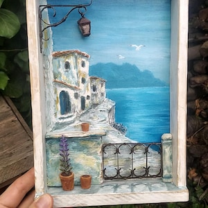 May include: A three-dimensional diorama in a white wooden frame depicting a coastal Mediterranean scene. The artwork features white buildings, a blue sea, a black metal lantern, a wrought iron railing, and small terracotta pots with plants.