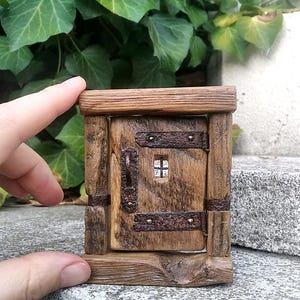 Könnte beinhalten: Ein kleiner, rechteckiger Türrahmen aus Holz mit einer zentralen Tür, die ein kleines, quadratisches Fenster besitzt. Die Struktur besteht aus dunklem, strukturiertem Holz und ist mit rostigen Metallscharnieren und einem Griff versehen.