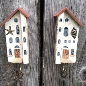 Puede incluir: Dos llaveros decorativos con forma de casitas, con tejados rojos y paredes blancas. Uno tiene una estrella de mar, el otro una concha. Ambos tienen una llave vintage colgando. El fondo es una superficie de madera envejecida. Aproximadamente 15 cm de altura.