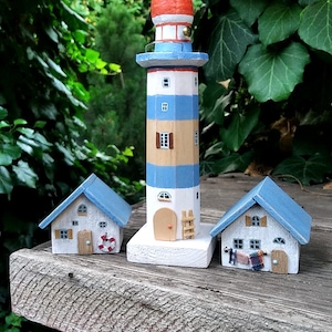 Puede incluir: Un faro de madera y dos casas en miniatura. El faro está pintado de blanco, azul y naranja, con una pequeña escalera. Las casas tienen tejados azules y paredes blancas. La escena está sobre una superficie de madera.