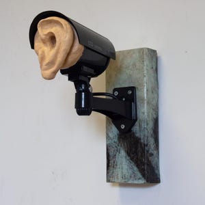 May include: A black CCTV camera with the text "CCD camera" is mounted on a weathered, rectangular block. A tan ear sculpture replaces the camera lens. The sculpture is attached to a wall.