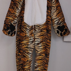 Puede incluir: Disfraz de tigre de cuerpo entero con capucha y cremallera. El patrón de rayas naranja y negro cubre todo el traje, con un vientre blanco y detalles de patas negras en manos y pies.