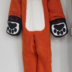 May include: Orange fox onesie with a hood, black paw-shaped mittens, and black feet. The onesie has a white belly and black accents on the ears. The costume is hanging on a white wall.