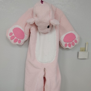 Puede incluir: Un disfraz de bebé de felpa rosa con forma de animal, colgado de una pared. El disfraz tiene una capucha con orejas, una barriga blanca y grandes almohadillas rosas en las manos y los pies. Una cremallera recorre la parte delantera.