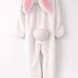 May include: A white, fluffy bunny onesie with a hood featuring pink-lined ears. The costume has a round, fluffy tail on the back and pink paw prints on the feet. The onesie is hanging against a white background.