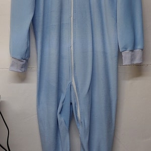 May include: Light blue one-piece pajama with a full-length zipper and footed bottoms. The collar and cuffs have a striped pattern. The pajamas are hanging on a white hanger against a white wall.