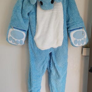 Puede incluir: Un mono de felpa azul claro con capucha con la cara de un animal de dibujos animados. El mono tiene una barriga blanca, detalles en forma de pata en las manos y cubiertas para los pies. El mono tiene una cremallera en las manos.