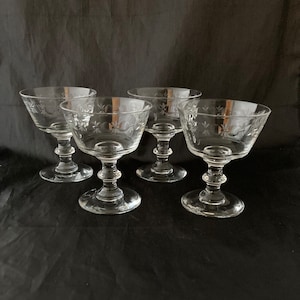 May include: Set of four clear glass champagne coupes with etched floral designs. The coupes have a delicate stem and a wide bowl.