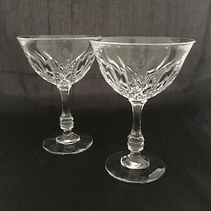 May include: Two clear glass champagne coupes with a cut glass design. The coupes have a tall stem and a wide bowl.