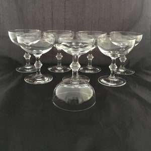 Peut inclure: Ensemble de huit coupes à champagne en verre transparent avec un large bol et une courte tige. Les coupes sont disposées en cercle sur une surface noire.