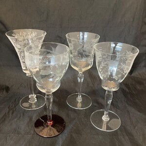 May include: Four clear etched glass goblets with floral designs. The goblets have varying bowl shapes and stem designs. One goblet has a dark red base. The glasses are arranged on a dark background.