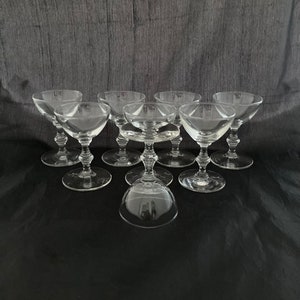 May include: A collection of eight clear, stemmed coupe glasses. The glasses have a shallow, rounded bowl and a delicate stem. The glasses are arranged on a dark surface, with one glass inverted in the foreground. The background is a dark gray.