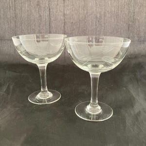 May include: Two clear glass champagne coupes with etched designs. The coupes are on a dark gray surface.