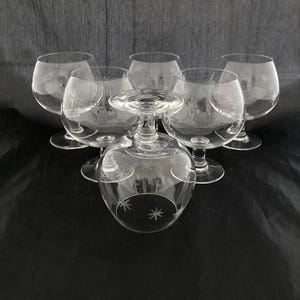 May include: Six clear brandy snifters with etched starburst designs. The glasses have a rounded bowl shape and a short stem. The glasses are arranged on a dark surface, with one glass inverted in the center.