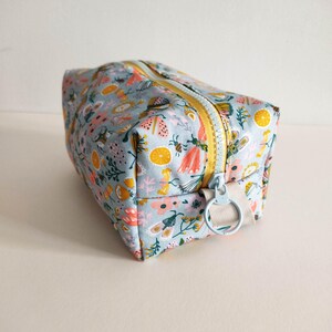 May include: A light blue zippered pouch with a floral print featuring butterflies, flowers, and lemons. The zipper has a yellow pull tab and a light blue ring.